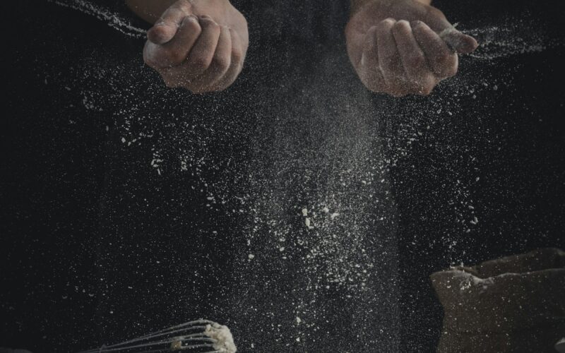 Flour falling onto a table beside eggs and a whisk