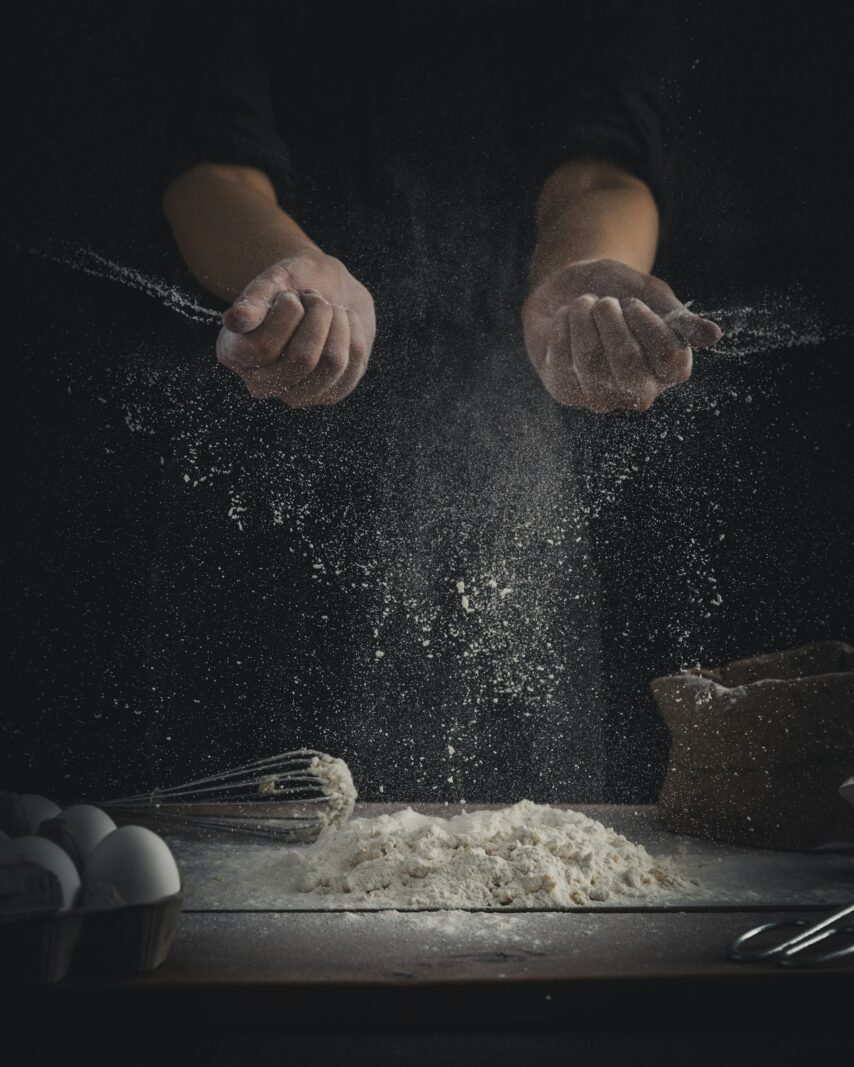 Flour falling onto a table beside eggs and a whisk